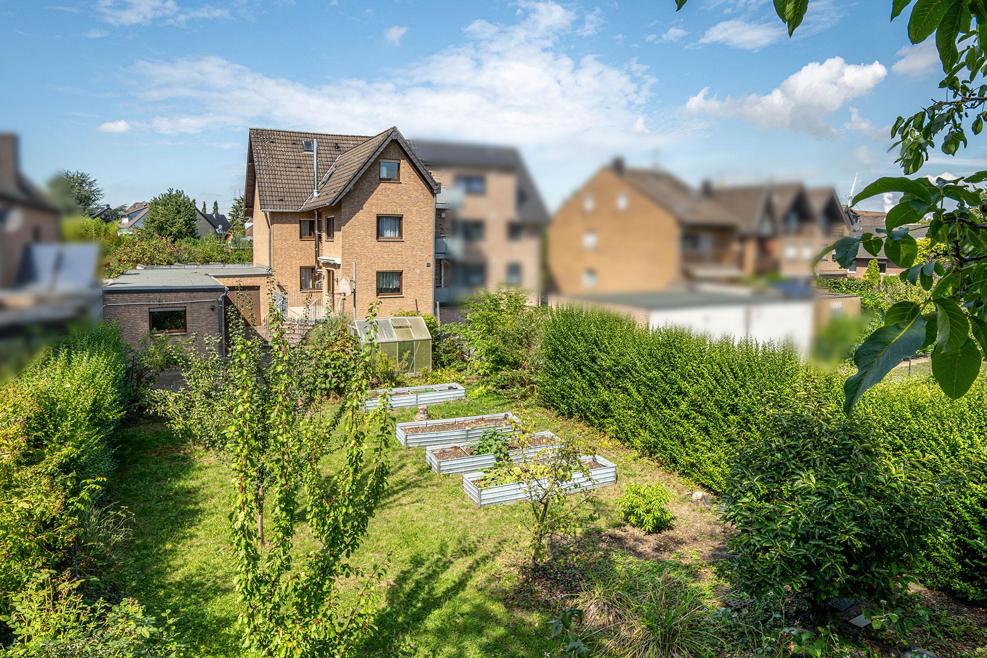 Das Bild zeigt den weitläufigen Garten und die Rückansicht der Immobilie. Neben dem Anbau an die Garage, der aus Büro, Werkraum oder Freizeitraum genutzt werden kann, befindet sich auch eine große gepflasterte Hoffläche direkt am Haus. Der große Garten ist mit Rasen, Hochbeeten und Obstbäumen gestaltet.Hier kann getobt und Obst und Gemüse für die selbstversorgung angebaut werden. Doppelhaushälfte Bergheim