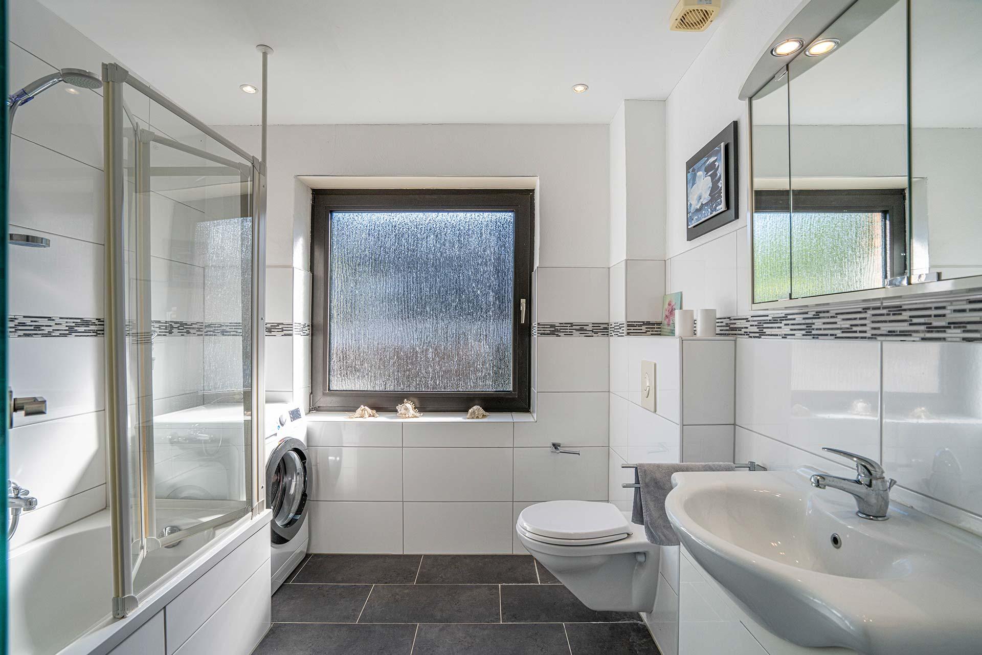 Das Wannenbad in Wohnung #2 ist mit einer Duschwanne, einem wandhängenden WC und einem Haundwaschbecken ausgestattet. Zudem befindet sich der Anschluss für eine Waschmaschine hier. Immobilie Bedburg